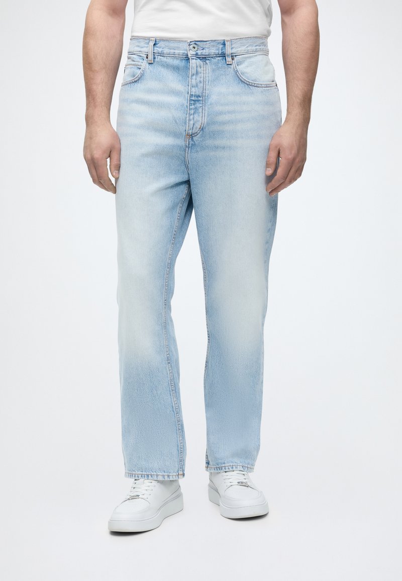 Homme portant un jean bleu clair droit et des baskets blanches, debout devant un fond blanc uni.