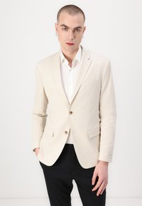 Lichtbeige textuurblazer met een enkele knoopsluiting, met opstaande revers en twee voorkeurszakken, gecombineerd met een wit shirt en zwarte broeken.