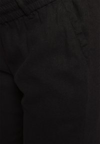 Gros plan sur un pantalon en tissu noir avec taille élastique et détail de poche latérale.