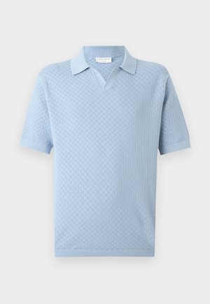 Jeune homme portant un polo en maille bleu clair à manches courtes, un pantalon slim gris et des baskets blanches, debout devant un fond uni.