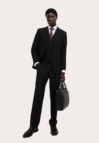 Schwarzer dreiteiliger Anzug mit weißem Hemd und roter Krawatte. Das Model hält eine schwarze Lederaktentasche und trägt glänzend schwarze Schuhe.