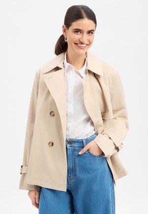 Woman smiling, wearing beige double-breasted trench coat over white shirt and blue jeans, with one hand in pocket, against white background.