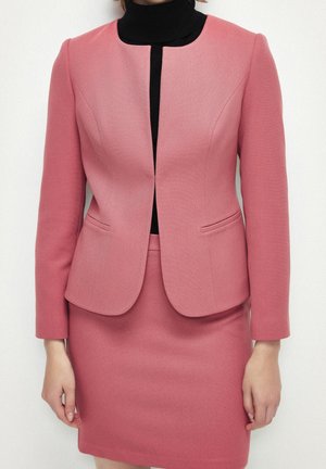 Kobieta ubrana w dopasowaną różową marynarkę i pasującą spódnicę, pod którą ma czarny golf, stoi na tle jednolitego białego tła.