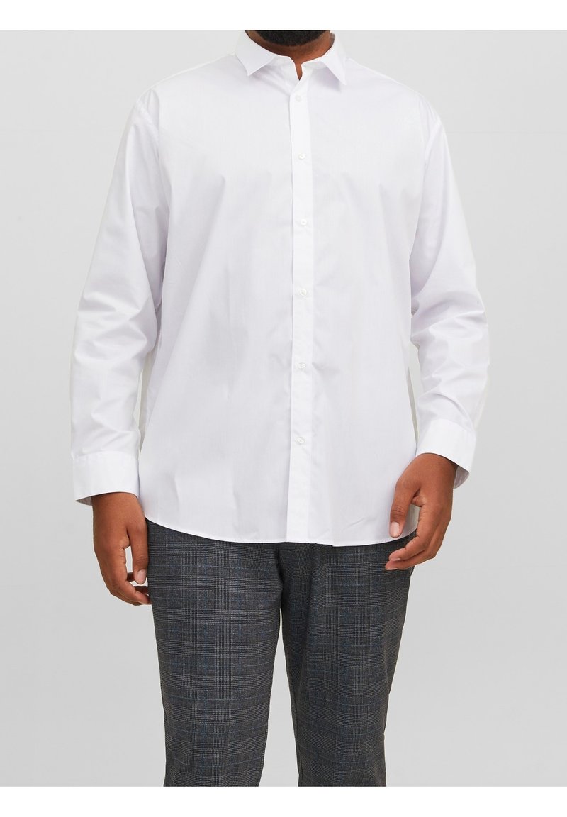 Homme portant une chemise blanche unie à manches longues et un pantalon à carreaux gris foncé, debout devant un fond gris clair.