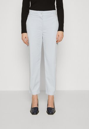 Pantalon bleu clair en tissu lisse, présentant une coupe droite et une fermeture à bouton. Porté avec des talons carrés sombres et brillants.