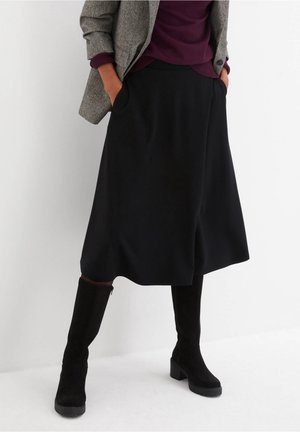Persona che indossa una gonna midi nera con tasche, stivali neri al ginocchio, un maglione viola e un blazer grigio in tweed con le mani nelle tasche.