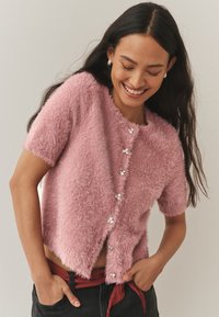 Rožinis pūkuotas trumpomis rankovėmis megztinis su apvalia iškirpte, puoštas dekoratyviniais sidabriniais mygtukais ir šiek tiek sutrumpintu dizainu.