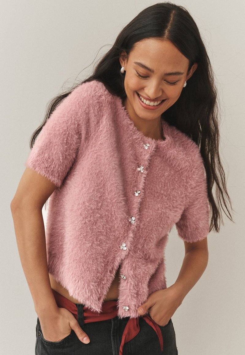 Rožinis pūkuotas trumpomis rankovėmis megztinis su apvalia iškirpte, puoštas dekoratyviniais sidabriniais mygtukais ir šiek tiek sutrumpintu dizainu.