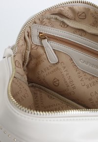 Interno della borsa color crema con tasca con zip, foderato in tessuto beige chiaro con motivo logo ripetuto e zip in tonalità oro.