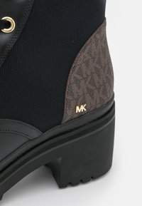 Bottine en cuir noir et maille avec un accent monogramme marron, un détail logo doré, et une semelle en caoutchouc texturée avec un talon bloc.