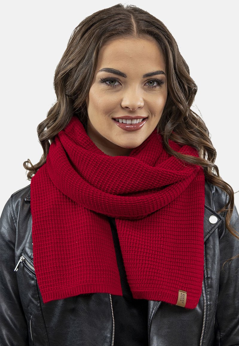Écharpe en tricot rouge avec un motif texturé, drapée élégamment autour du cou, assortie à une veste en cuir noire et avec une étiquette sur l'écharpe.