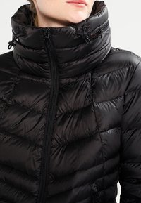 Musta puhallettava takki korkealla kauluksella, tikattu rakenne ja vetoketjusuljenta. Varusteena narut kauluksessa säädettävyyttä varten.