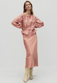 Różowa bluzka z kwiatowym wzorem i wiązaniem z przodu, w połączeniu z satynową, różową spódnicą. Modelka ma na sobie kremowe kozaki za kolano.