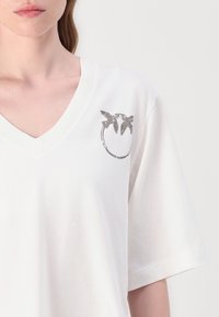 T-shirt in cotone bianco con scollatura a V, caratterizzato da una grafica argento di uccelli all'interno di un cerchio nella zona sinistra del petto. Maniche corte.