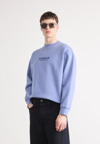 Világoskék pulóver kerek nyakkal és hosszú ujjal, a "BARBOUR INTERNATIONAL" sötét nyomott logóval. Sötét farmernadrággal viselve.