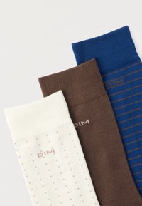 Τρία διπλωμένα καλτσάκια, ένα λευκό με μικρές βούλες, ένα μονόχρωμο καφέ, ένα μπλε με λεπτές καφέ ρίγες, όλα με την επωνυμία "DIM" κοντά στις μανσέτες.