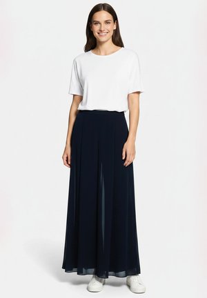 Femme souriante aux longs cheveux foncés portant un haut blanc à manches courtes, une longue jupe bleu marine et des baskets blanches, debout devant un fond uni.