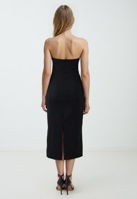 Schwarzes, trägerloses Midikleid mit tailliertem Oberteil, seitlichen Nähten und einem Schlitz im Rücken. Getragen mit schwarzen High Heels.