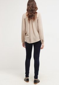 Blouse beige à manches longues avec une coupe ample, présentant un ourlet droit à l'arrière, des poignets boutonnés et un tissu doux et lisse. Porté avec un jean skinny noir.