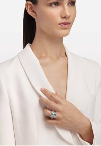 Srebrni prstan z več plastmi, ki vključuje gravirane akcentne dele in svetlo modro emajlirano vstavitev. Model nosi bel blazer.