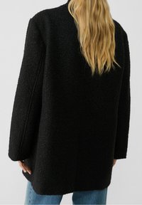 Abrigo negro texturizado con corte oversize, que presenta mangas amplias y un diseño de espalda recta. Estilo casual combinado con jeans azul claro.