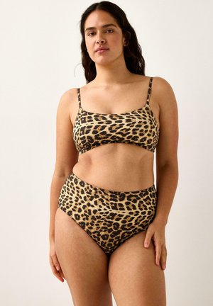 Donna in piedi davanti a uno sfondo neutro, indossa un bikini con stampa leopardata con top a scollo quadrato e slip a vita alta, rivolta verso l'obiettivo.