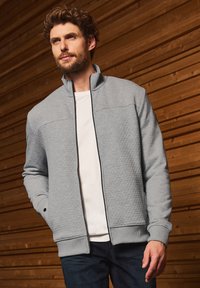 Veste zippée gris clair avec une surface texturée, col et poignets côtelés, dotée de poches latérales et d'une fermeture éclair noire contrastante.