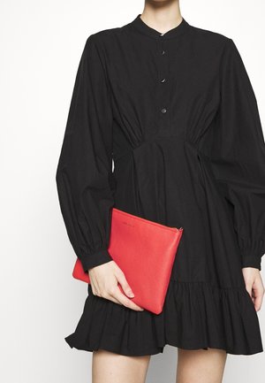 Clutch in pelle rossa con finitura testurizzata, chiusura a zip e logo in rilievo, tenuta accanto a un vestito nero con maniche a sbuffo e orlo arricciato.