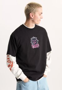 Camiseta gráfica de algodón negro con un logo circular con púas en rosa y negro. Superpuesta con una camisa de manga larga blanca debajo.