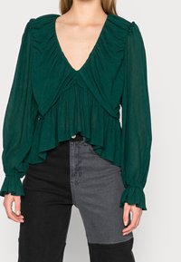 Blusa verde smeraldo con scollatura arricciata e maniche lunghe, caratterizzata da un tessuto texturizzato e un orlo a strati. Abbinata a jeans neri e grigi.