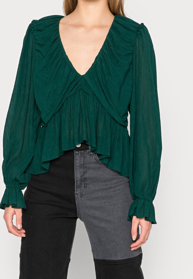 Blusa verde smeraldo con scollatura arricciata e maniche lunghe, caratterizzata da un tessuto texturizzato e un orlo a strati. Abbinata a jeans neri e grigi.