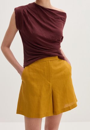 Femme portant un top asymétrique sans manches bordeaux rentré dans un short taille haute large jaune moutarde, mains dans les poches.