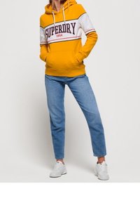 Sudadera amarilla con rayas horizontales blancas y rojas, con el logo "SUPERDRY". Combinada con jeans azul claro y zapatillas blancas.