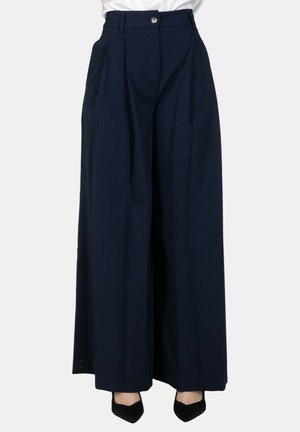 Donna che indossa pantaloni blu navy a vita alta e gamba larga abbinati a décolleté nere con punta appuntita su sfondo bianco semplice.