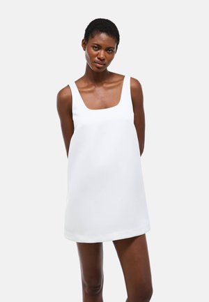 Femme aux cheveux courts portant une robe blanche sans manches avec un col carré, debout devant un fond clair uni.