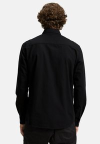 Camisa negra de manga larga con cuello abotonado, de corte recto y con una tela texturizada. Presenta puños abotonados y un detalle en el yugo trasero.