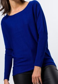 Haut en maille bleu à manches longues avec un large col, doté d'une coupe décontractée et de poignets côtelés, associé à un pantalon noir ajusté.