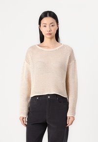 Beige cropped sweater with a loose, knitted texture and wide crew neckline. Paired with black pants, it has long sleeves and ribbed cuffs.
