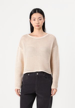 Pull cropped beige avec une texture tricotée lâche et un large col rond. Associé à un pantalon noir, il possède des manches longues et des poignets côtelés.