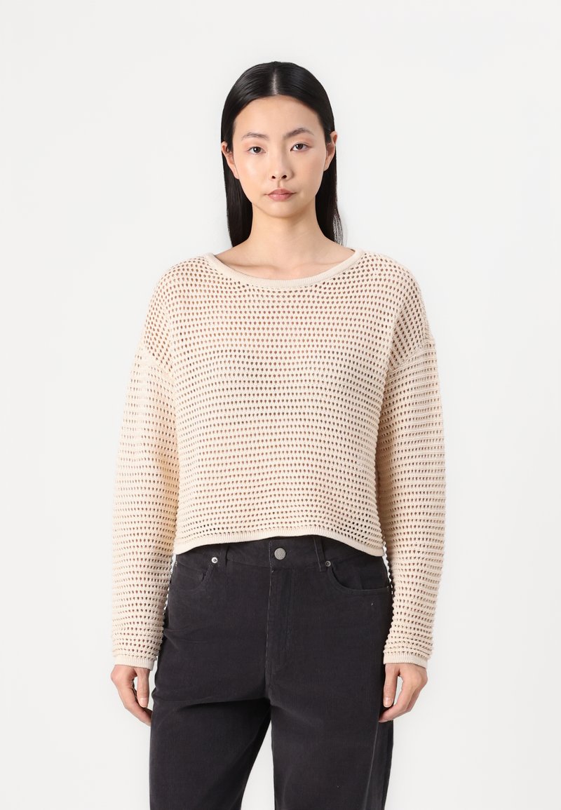 Beige cropped sweater with a loose, knitted texture and wide crew neckline. Paired with black pants, it has long sleeves and ribbed cuffs.