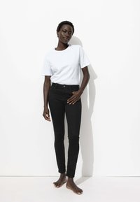 Jean noir skinny avec cinq poches et fermeture à bouton, associé à un t-shirt en coton blanc uni à manches courtes, sur un modèle pieds nus.