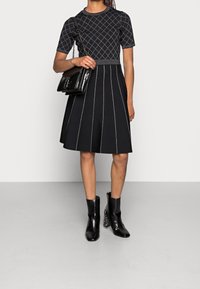 Robe noire à manches courtes avec motif en losanges et coutures blanches en accent, jupe évasée. Assortie à des bottines noires et un sac à main noir brillant.
