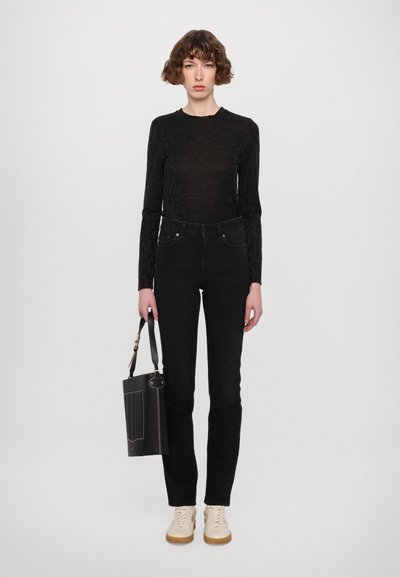 Top negro de manga larga con un patrón en relieve tonal, combinado con jeans negros de cintura alta y zapatos beige. Se sostiene un pequeño bolso negro.