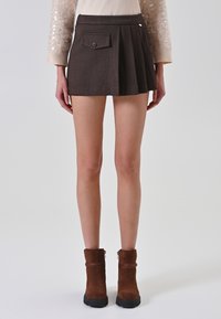 Donna che indossa una mini gonna plissettata color marrone scuro con una tasca a bottoni, un maglione beige e stivaletti marroni su uno sfondo chiaro.