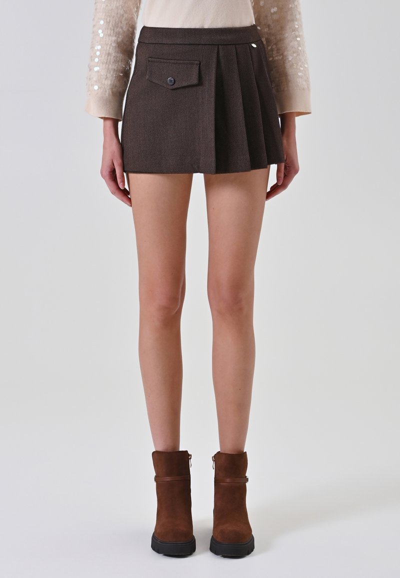 Donna che indossa una mini gonna plissettata color marrone scuro con una tasca a bottoni, un maglione beige e stivaletti marroni su uno sfondo chiaro.