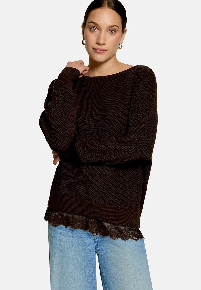 Jeune femme aux cheveux foncés portant des boucles d'oreilles créoles en or, un pull ample marron foncé, un haut orné de dentelle et un jean bleu clair, debout les bras croisés.