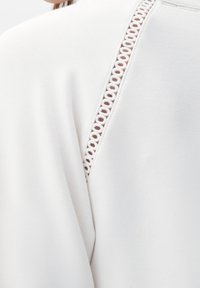White sweatshirt featuring a textured fabric with a curved seam and decorative woven details along the back seam.
