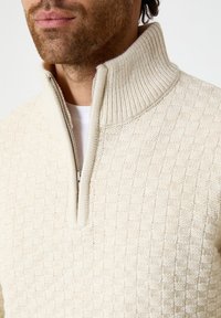 Pull à zip crème avec col montant, présentant un motif en losange texturé et des accents côtelés au niveau du col et de la fermeture éclair.