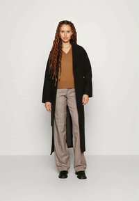Woman with long curly hair wearing a black long coat, brown V-neck sweater, plaid wide-leg pants, and black shoes, standing against a plain background.