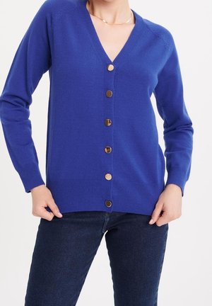 Frau trägt eine königsblaue V-Ausschnitt-Strickjacke mit sieben goldenen Knöpfen, kombiniert mit dunkelblauen Jeans, Hände nahe den Taschen ruhend.